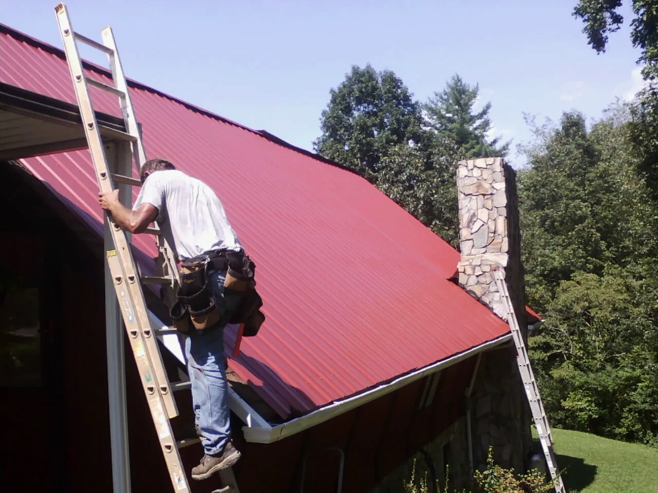 Roofer on a ladder beside a red metal roof and stone chimney — Soffit & Fascia Repair by Roofing Brothers in Tunica